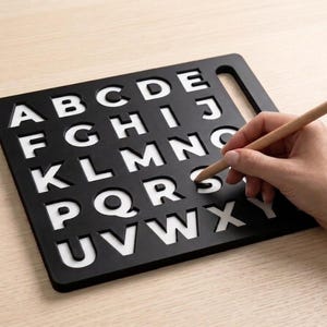 May include: A black rectangular alphabet stencil board featuring cut-out capital letters from A to Y arranged in five rows. A light-colored wooden stylus is positioned to trace the letter S within the stencil, which rests on a flat surface.