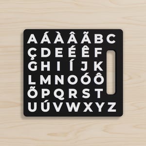 May include: A black alphabet board with white uppercase letters, including accented characters. The board has a rectangular shape with a handle cutout on the right side. The letters are arranged in rows, with each letter clearly visible against the black background.
