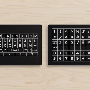 May include: Two black rectangular keyboard layouts with white lettering and symbols. One features a QWERTY keyboard, the other includes numbers, symbols, and functions. The layouts are on a light wood grain surface.