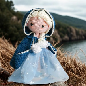 Pu&ograve; includere: Bambola all'uncinetto fatta a mano, vestita con un mantello blu con cappuccio e un vestito azzurro. La bambola ha i capelli biondi e si trova in un ambiente naturale all'aperto, con uno specchio d'acqua sullo sfondo. L'abito della bambola ha un rivestimento trasparente.