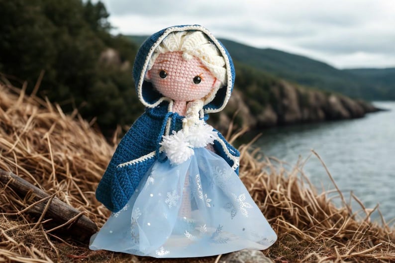 Pu&ograve; includere: Bambola all'uncinetto fatta a mano, vestita con un mantello blu con cappuccio e un vestito azzurro. La bambola ha i capelli biondi e si trova in un ambiente naturale all'aperto, con uno specchio d'acqua sullo sfondo. L'abito della bambola ha un rivestimento trasparente.