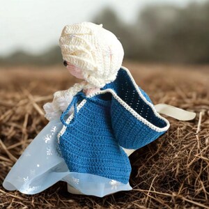 Pu&ograve; includere: Bambola all'uncinetto fatta a mano con un mantello blu con finiture bianche e un cappuccio bianco. La bambola ha un viso rosa chiaro ed &egrave; ambientata su uno sfondo sfocato di erba secca.