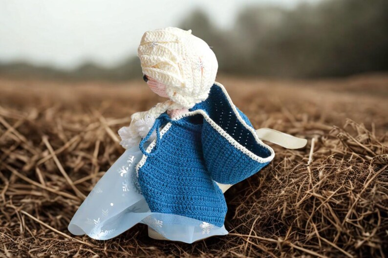 Pu&ograve; includere: Bambola all'uncinetto fatta a mano con un mantello blu con finiture bianche e un cappuccio bianco. La bambola ha un viso rosa chiaro ed &egrave; ambientata su uno sfondo sfocato di erba secca.