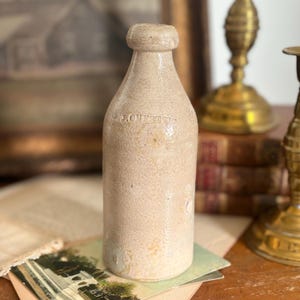 May include: A vintage, beige-colored bottle with a rounded top and a cylindrical body. The bottle has a textured surface and appears to be made of stoneware. The text "J. OUTTER" is visible on the bottle. The bottle is displayed on a table with other antique items.