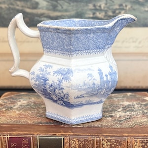May include: Antique blue and white ceramic pitcher with a detailed landscape design. The pitcher features a curved handle and spout, with a decorative pattern around the rim and base. The overall design is reminiscent of vintage tableware.