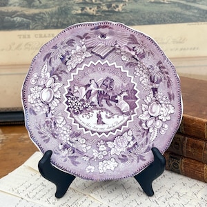 May include: A purple antique plate with a detailed design, featuring a central scene with figures and text that reads "Peace on Earth" and "Give us this day our daily bread." The plate is supported by a black stand.