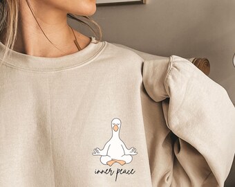 Könnte beinhalten: Beiges Sweatshirt mit einer Cartoon-Gans in einer Yoga-Pose und dem Schriftzug "inner peace". Die Gans ist weiß mit gelbem Schnabel, sitzt im Schneidersitz. Das Sweatshirt hat einen Rundhalsausschnitt und lange Ärmel. Der Text ist in schwarzer Schrift.
