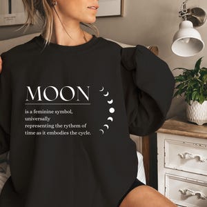 Könnte beinhalten: Schwarzer Pullover mit weißem Text, der "MOON is a feminine symbol, universally representing the rhythm of time as it embodies the cycle." lautet. Der Text ist von einer Reihe weißer Halbmonde umgeben.