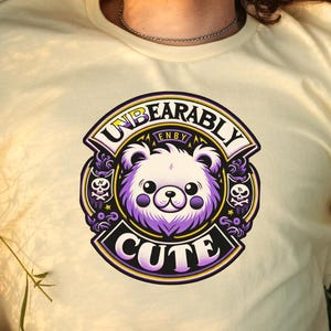 Pode incluir: Uma pessoa vestindo uma camiseta amarela com um gráfico roxo e amarelo de um urso de desenho animado com o texto "Unbearably Cute" em um círculo.
