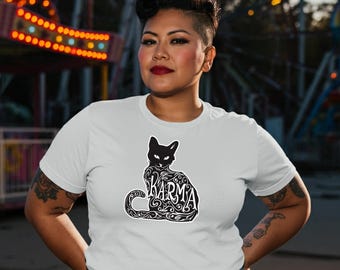 Könnte beinhalten: Weißes T-Shirt mit einem schwarzen Katzen-Design und dem Wort "KARMA" in einer dekorativen Schriftart. Die Katzenillustration ist mit Wirbelmustern versehen. Die Person trägt eine blaue Jeans. Der Hintergrund zeigt einen verschwommenen Vergnügungspark.