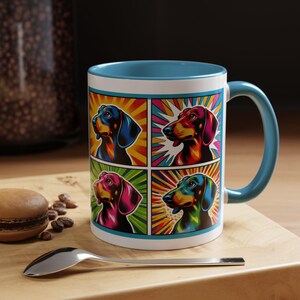 Może przedstawiać: Biały kubek ceramiczny z niebieskim rantem i uchwytem. Kubek przedstawia kolorowy projekt w stylu pop art z czterema psami rasy dachshund na jasnym żółtym, czerwonym, zielonym i niebieskim tle.