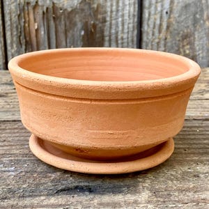 May include: A small, round terracotta pot with a matching saucer. The pot has a wide, shallow bowl shape with a slightly textured surface and a warm, earthy tone. Ideal for small plants or succulents.