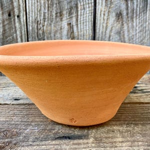 May include: A terracotta-coloured, shallow bowl with a wide, flared rim. The bowl has a smooth, unglazed surface and a gently curved shape. The bowl is likely used for planting or decorative purposes.