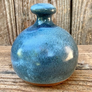 May include: A small, round ceramic vase with a narrow neck and a flared rim. The body of the vase features a glossy, mottled glaze in shades of deep blue, teal, and light blue, while the base reveals the natural, unglazed reddish-brown clay.