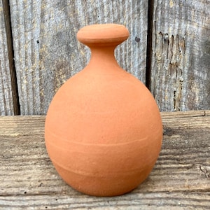 Op de afbeelding: Een terracotta-kleurige vaas met een rond lichaam, een smalle hals en een platte, schijfvormige bovenkant. De vaas heeft een glad oppervlak met een subtiele horizontale groef in het midden. De achtergrond is een verweerd houten oppervlak.