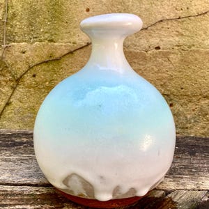 May include: A small, round ceramic vase with a narrow neck and a flared rim. The body features a smooth, glossy glaze that transitions from a soft, pale blue at the top to a creamy white at the base, where the glaze drips over a terracotta bottom.