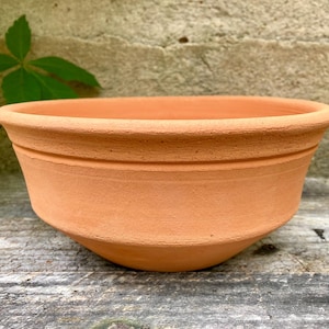 May include: A terracotta-colored, round plant pot with a wide, shallow bowl shape. The pot has a slightly textured surface and a decorative band near the rim. The pot sits on a weathered wooden surface.