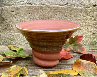 May include: A terracotta-coloured ceramic bowl with a wide, slightly flared rim. The bowl has a spiral-shaped base and a glazed, brown and green exterior. The bowl is surrounded by colourful autumn leaves on a wooden surface.