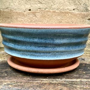 May include: A round terracotta planter with a wide, flared rim and horizontal ribbed texture. The upper portion features a speckled, light blue glaze that fades into the natural reddish-brown clay base, accompanied by a matching circular saucer.
