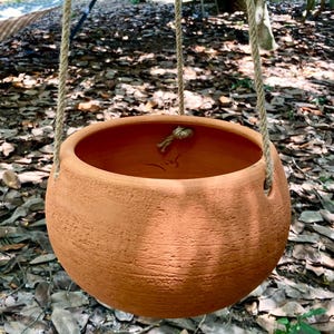 May include: A round, terracotta-colored clay hanging planter with a textured, matte surface. The bowl-shaped vessel features three evenly spaced holes along the upper rim, threaded with thick, natural-colored jute rope for suspension.