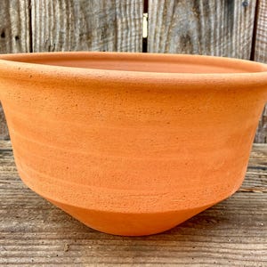 May include: A terracotta-colored, unglazed ceramic planter with a wide, rounded bowl shape. The planter has a slightly flared rim and a textured surface. The exterior is a warm, earthy orange hue, suitable for various plants and garden settings.