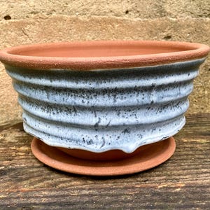 May include: A terracotta planter with a ribbed exterior and a matching saucer. The upper portion of the planter features a white glaze with dark gray speckles that drips down the sides, leaving the natural reddish-brown clay visible at the base.