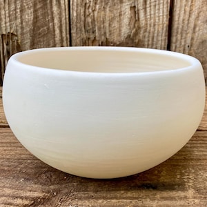 May include: A round, off-white ceramic bowl with a smooth, matte finish. The bowl has a wide opening and a rounded base. The background is a wooden surface.