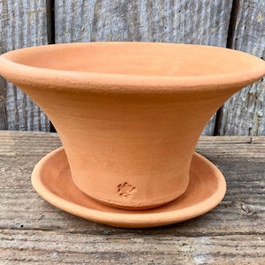 May include: A terracotta-colored, flared plant pot with a matching saucer. The pot has a wide, rounded rim and a slightly textured surface. The saucer is round and fits snugly beneath the pot. A small embossed design is visible on the saucer.