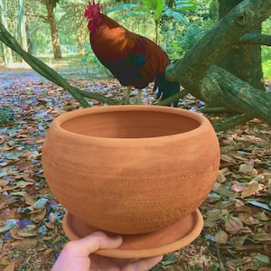 May include: A round, terracotta-colored clay planter pot with a matching circular saucer base. The pot features a textured, matte surface with visible horizontal striations and a wide, open top rim, designed for holding plants or decorative items.