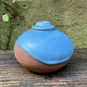 May include: A handcrafted ceramic vase with a rounded shape. The top half is a vibrant blue, contrasting with the earthy brown base. The vase has a small, raised rim at the top, adding a touch of elegance. Perfect for displaying flowers or as a decorative piece.