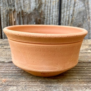 May include: A terracotta-colored, round, unglazed clay pot. The pot has a wide, shallow bowl shape with a slightly flared rim and a rounded base. The exterior has a textured surface, and the pot appears to be handmade.