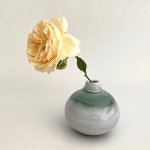 以下が含まれることがあります： 小さな丸い陶器の花瓶に、桃色のバラが活けられています。花瓶は、下部の淡いグレーから上部の柔らかな緑へとグラデーションになっています。バラは満開で、緑色の茎と葉が付いています。