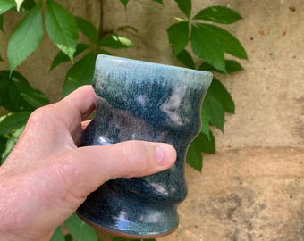 May include: A handmade ceramic mug with a unique, wavy design. The mug features a blend of teal and dark blue hues, with a lighter blue rim. The base has a reddish-brown color. The mug is held in a hand, with green leaves in the background.