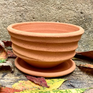 May include: A terracotta plant pot with a wide, circular opening and a matching round saucer. The exterior of the pot features a series of horizontal, raised ridges that wrap around the body, creating a textured, ribbed appearance in a natural clay color.
