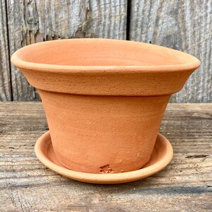 Może przedstawiać: Terakotowy, nieszkliwiony ceramiczny doniczka z pasującym spodkiem. Doniczka ma szeroką, lekko rozszerzoną krawędź i cylindryczny korpus. Spodek jest okrągły i idealnie pasuje pod spód, idealny do roślin.