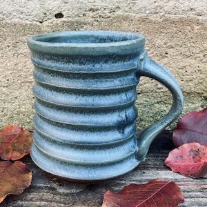 May include: A handmade, blue-green ceramic mug with a ribbed design. The mug has a curved handle and sits on a wooden surface with fallen red leaves. The mug is approximately 4 inches tall.