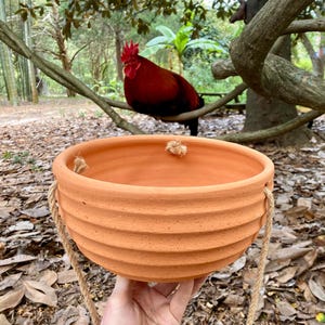 May include: A round, terracotta-colored hanging planter with a ribbed, horizontal texture on the exterior. The bowl features three evenly spaced holes along the upper rim, threaded with thick, natural-colored jute rope for suspension.