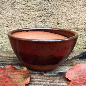 Op de afbeelding: Een handgemaakte keramische kom met een bruine glazuur. De kom heeft een afgeronde vorm met een donkere rand en een lichtere binnenkant. Hij staat op een houten oppervlak, vergezeld van gevallen bladeren.