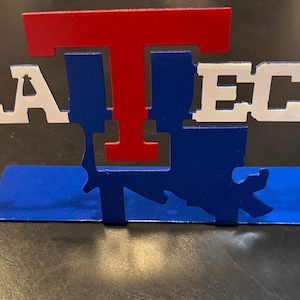 May include: A metal desk sign featuring the text LA TECH in white letters with a large red T centered above a blue silhouette of the state of Louisiana. The entire structure is supported by a flat blue rectangular base for stability.