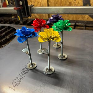 May include: Five metal roses in different colors, including red, blue, yellow, green, and purple, are arranged on a metal surface. The roses are made of metal and have a stem and a base.