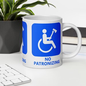 Puede incluir: Taza de cerámica blanca con un gráfico cuadrado azul. El gráfico representa a una persona en silla de ruedas sosteniendo un hacha. El texto "NO PATRONIZING" está impreso debajo del gráfico.