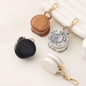 May include: Four small, round coin purses with gold-tone zippers and keychains. One is black, one is tan, one is white, and one is white with a black and white floral pattern. The purses are made of leather-like material.