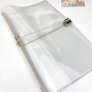 以下が含まれることがあります： 透明なプラスチック製のノートカバーで、柔軟なベージュのゴムバンドとシルバーの金属製留め具が付いています。カバーは角が丸い長方形で、ゴム紐を使って中身をしっかりと固定できるように設計されています。