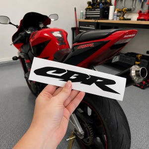 May include: A rectangular white decal featuring the black CBR logo in a stylized font. The sticker is held in front of a red Honda CBR motorcycle with a black seat, a black exhaust pipe, and a black rear tire, set against a garage background.