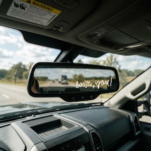 May include: A black rearview mirror inside a vehicle featuring the white cursive text "love you!" with a small heart symbol applied to the glass surface. The mirror reflects a road scene with a truck in the distance under a bright sky.