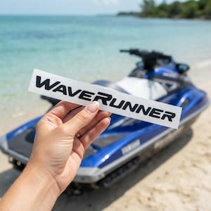 May include: A rectangular white decal featuring the black text "WAVERUNNER" in a bold, sans-serif font. The sticker is held horizontally, displaying the brand name clearly against a background that includes a blue personal watercraft.