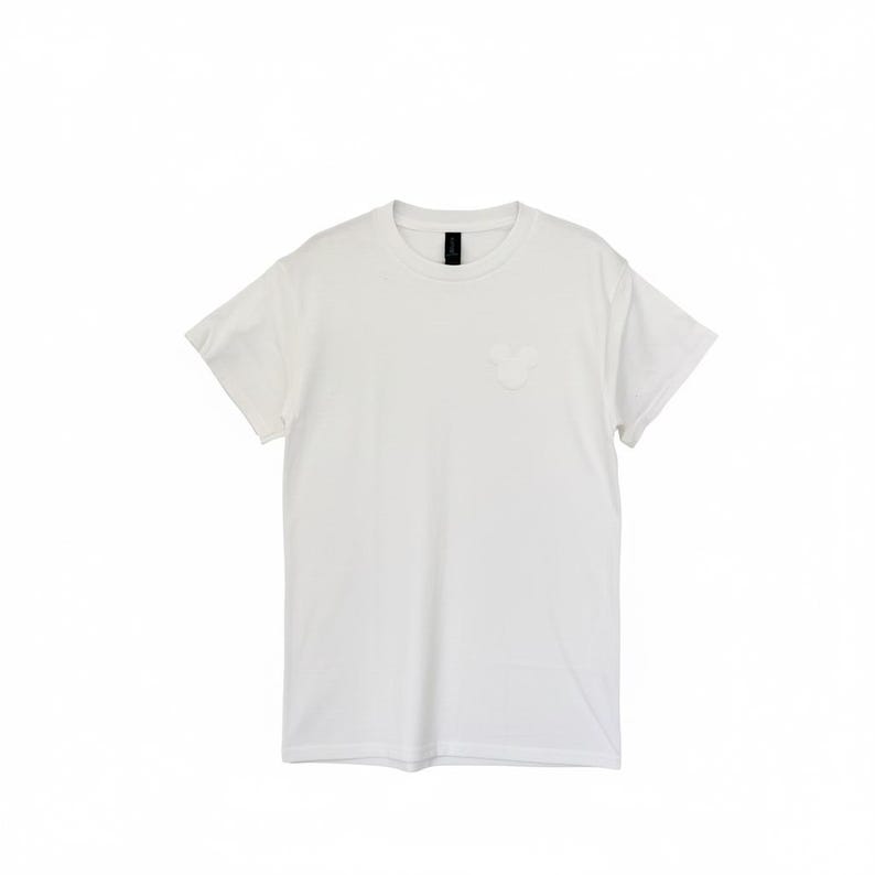 Puede incluir: Una camiseta blanca lisa con una peque&ntilde;a cabeza de Mickey Mouse en relieve en el pecho. La camiseta de manga corta tiene cuello redondo y un dise&ntilde;o sencillo. La tela parece suave y opaca.