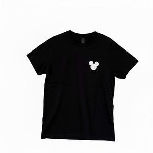 Puede incluir: Camiseta negra de cuello redondo con un logotipo blanco de la cabeza de Mickey Mouse en la parte superior izquierda del pecho. La camiseta es de material suave.
