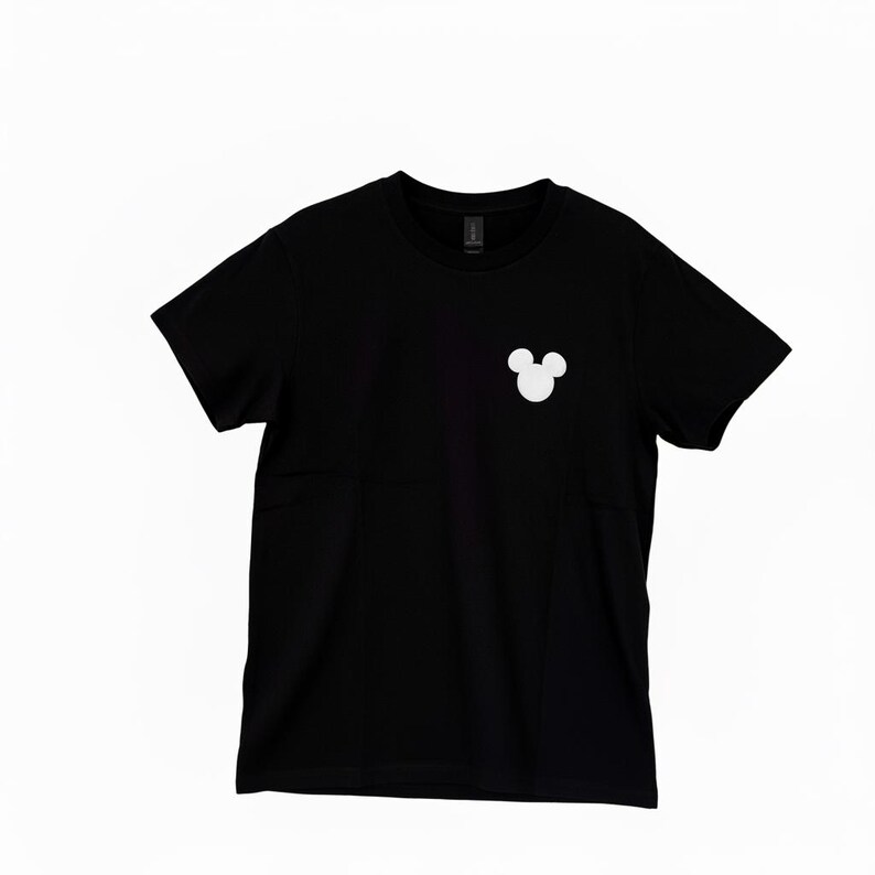 Puede incluir: Camiseta negra de cuello redondo con un logotipo blanco de la cabeza de Mickey Mouse en la parte superior izquierda del pecho. La camiseta es de material suave.