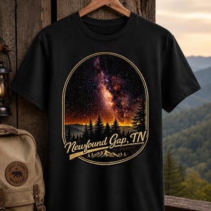 May include: A black short-sleeved t-shirt featuring a graphic print of a starry night sky over a forest silhouette. The design is enclosed in a gold-outlined arch and includes the text Newfound Gap, TN in a gold script font above a mountain icon.